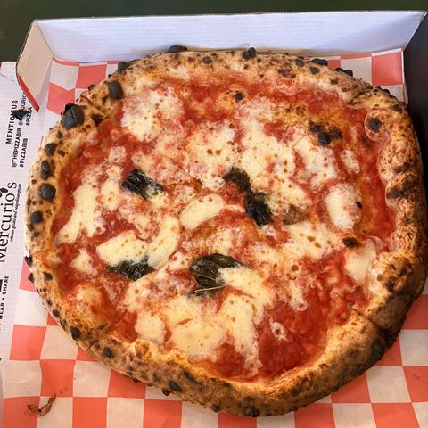 A wood-fired margherita pizza with charred crust and basil, in a takeout box from Mercurio's on red-and-white checkered paper